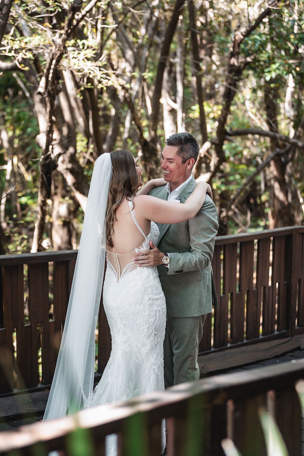 Bride in lace gown and veil embraces groom in sage suit on wooden deck surrounded by lush greenery.