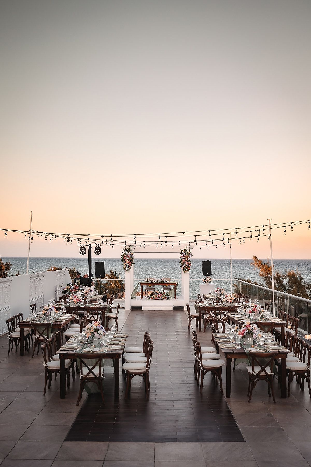 Elegant outdoor wedding reception setup on a seaside terrace at sunset with string lights, floral arrangements, and wooden chairs.