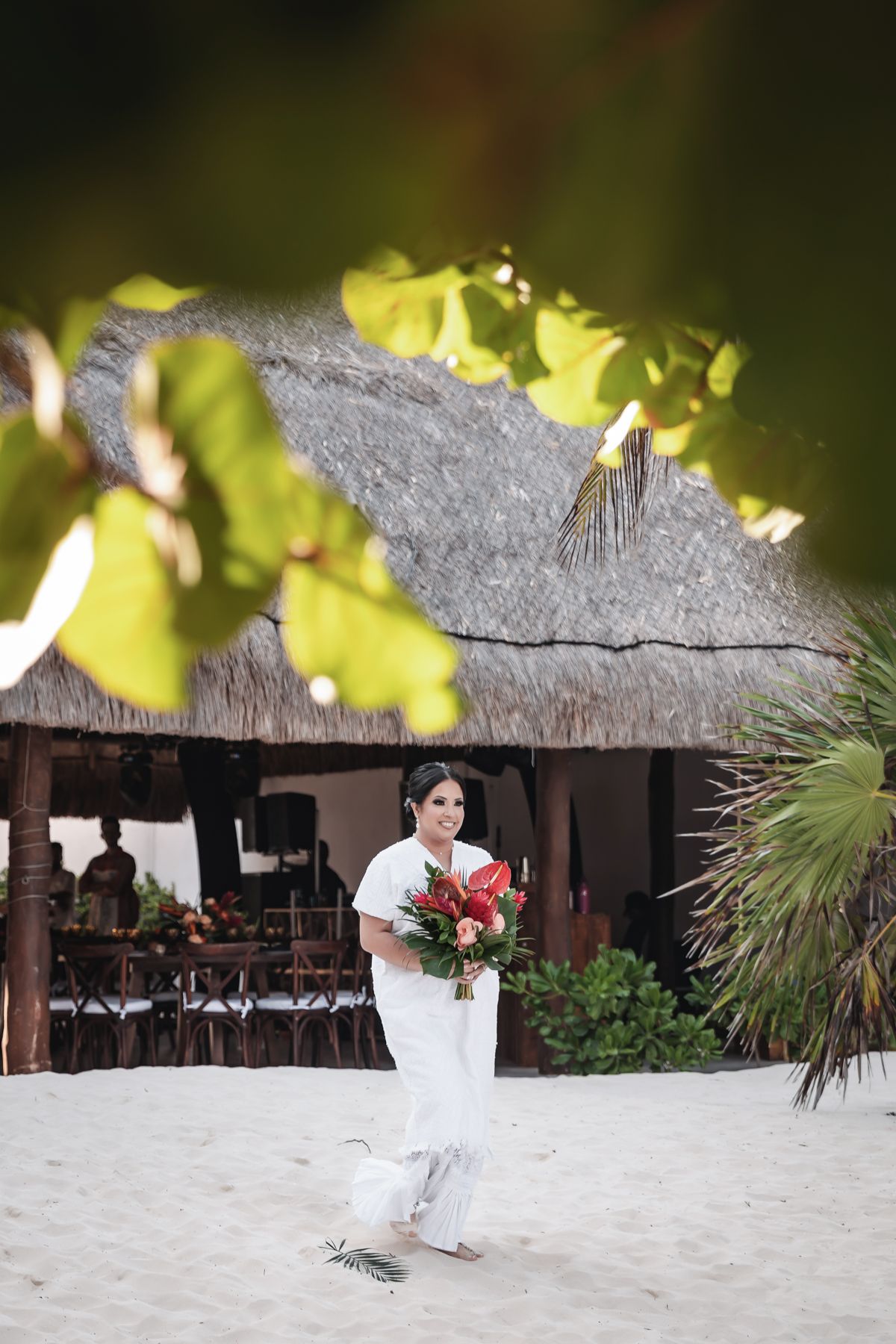 Bride in white dress holds tropical bouquet on sandy beach near thatched palapa at destination wedding.