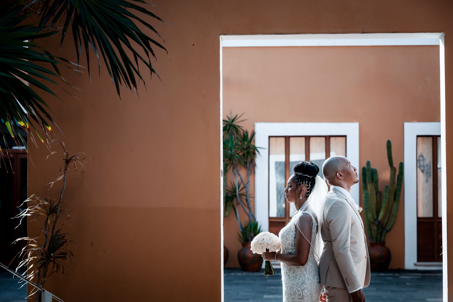 A bride and groom share a tender moment outside a terracotta building, bride holding white bouquet.