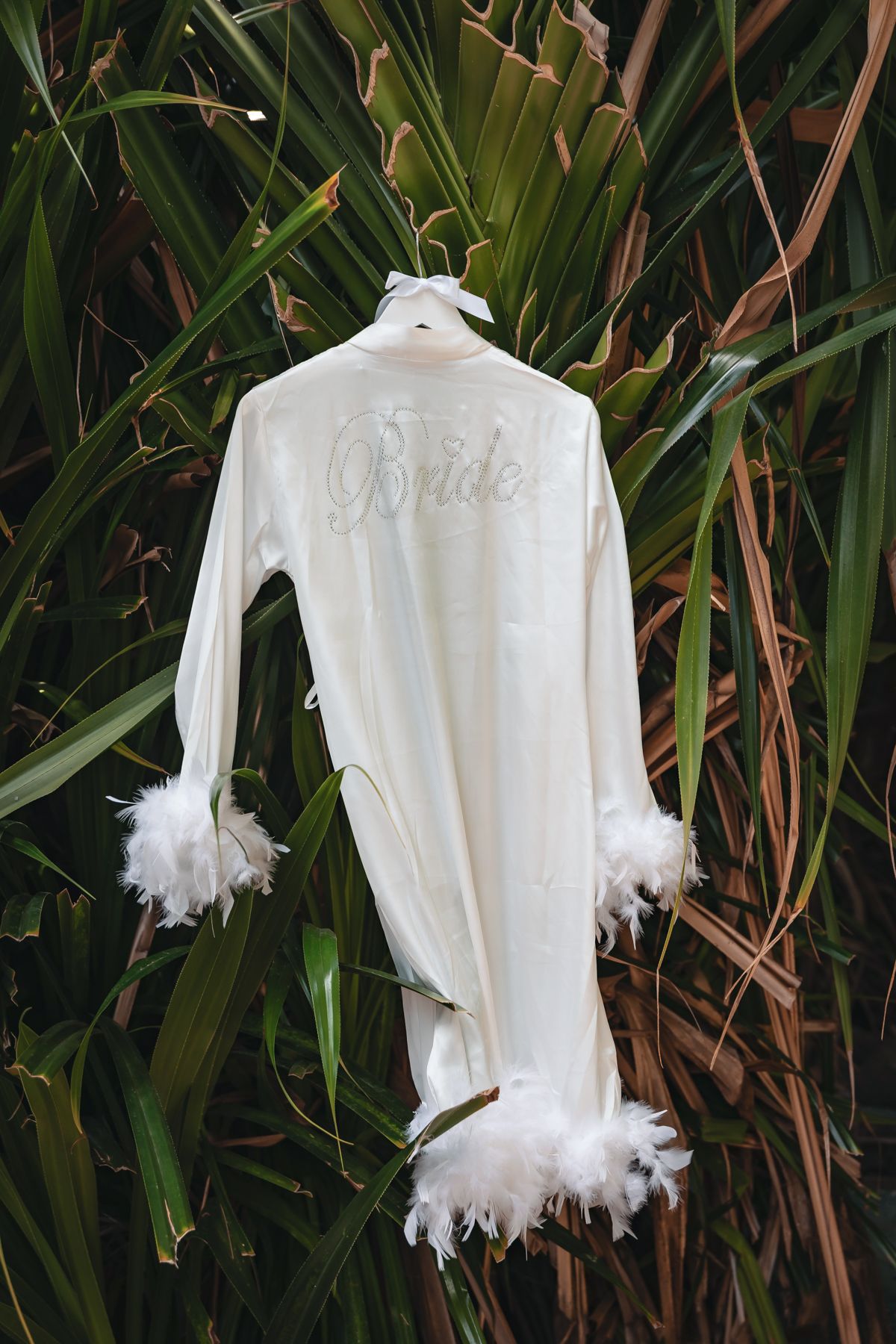 White feathered bridal robe with Bride embroidery hanging on tropical green foliage backdrop.
