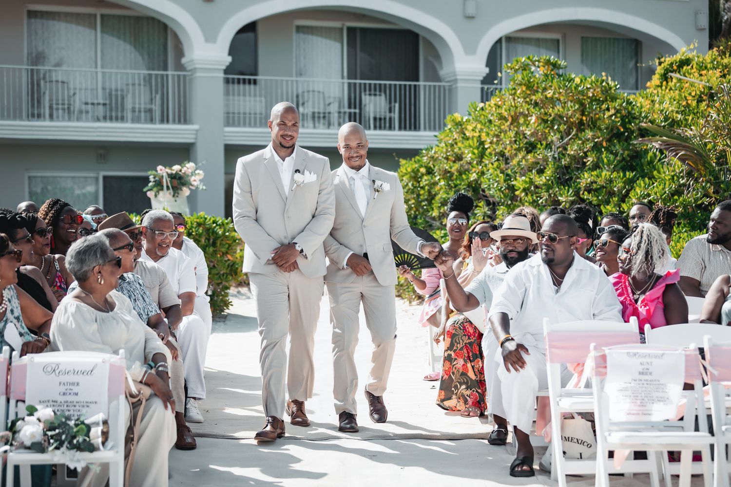 Two grooms in beige suits walk down the aisle at an outdoor wedding ceremony with smiling guests seated on both sides.