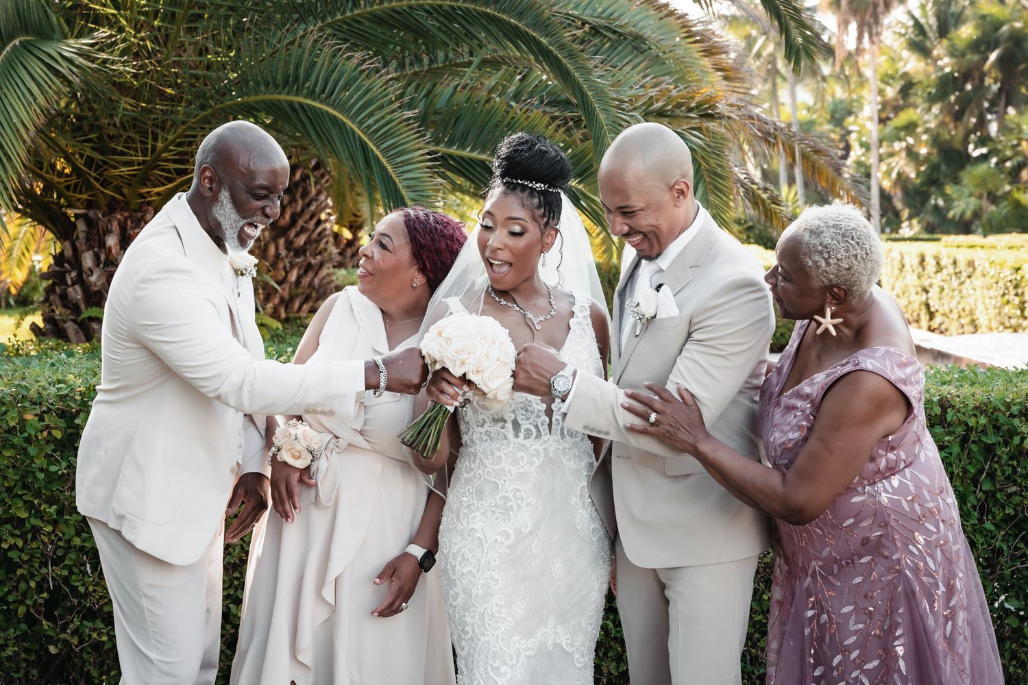 A joyful wedding party with bride and groom in white celebrating with family outdoors near tropical palm trees.
