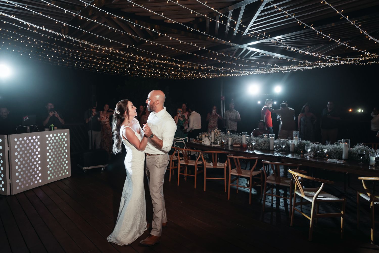 Bride and groom share first dance under twinkling fairy lights at elegant outdoor wedding reception at night.