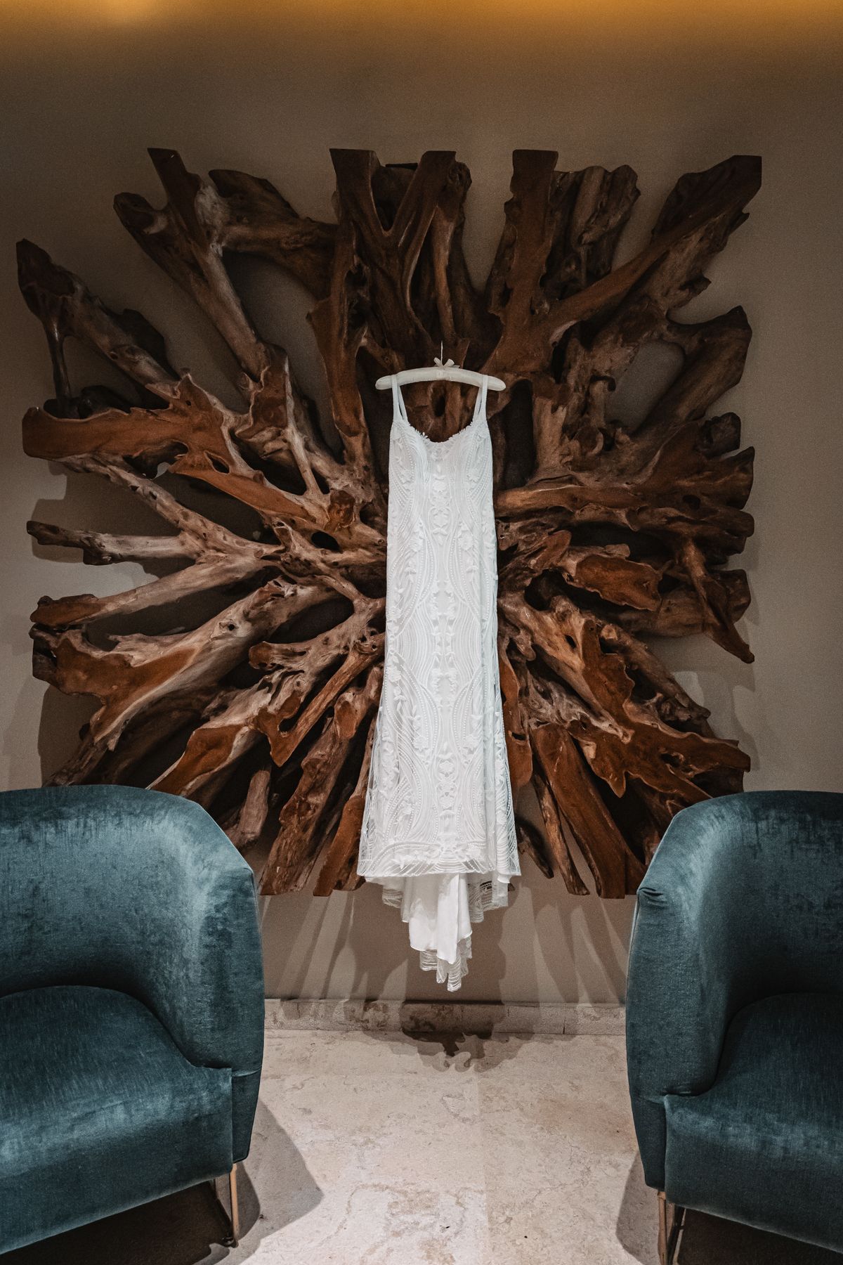 White lace wedding dress hanging on wooden art installation between two teal velvet chairs in elegant bridal suite.