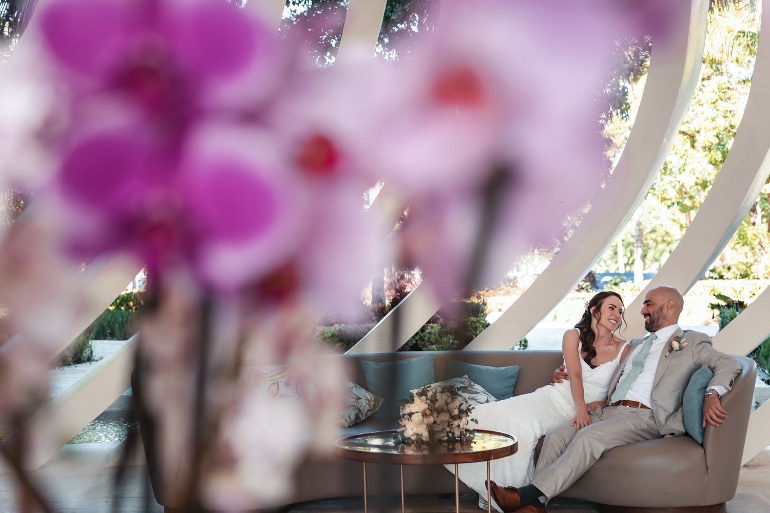Bride and groom relaxing on curved sofa surrounded by vibrant pink orchids in elegant venue.