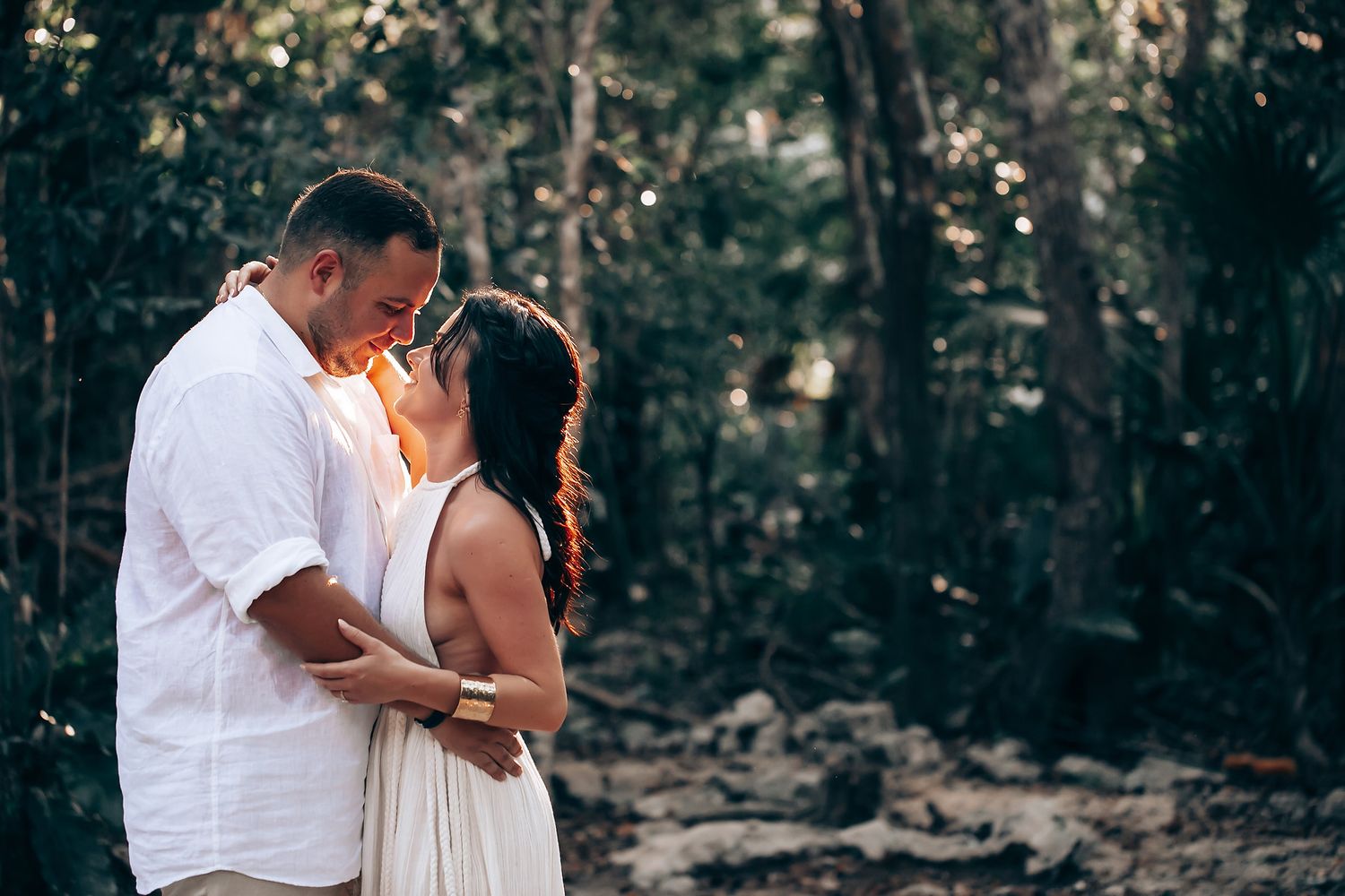 Intimate moment captured in white attire against lush forest backdrop.