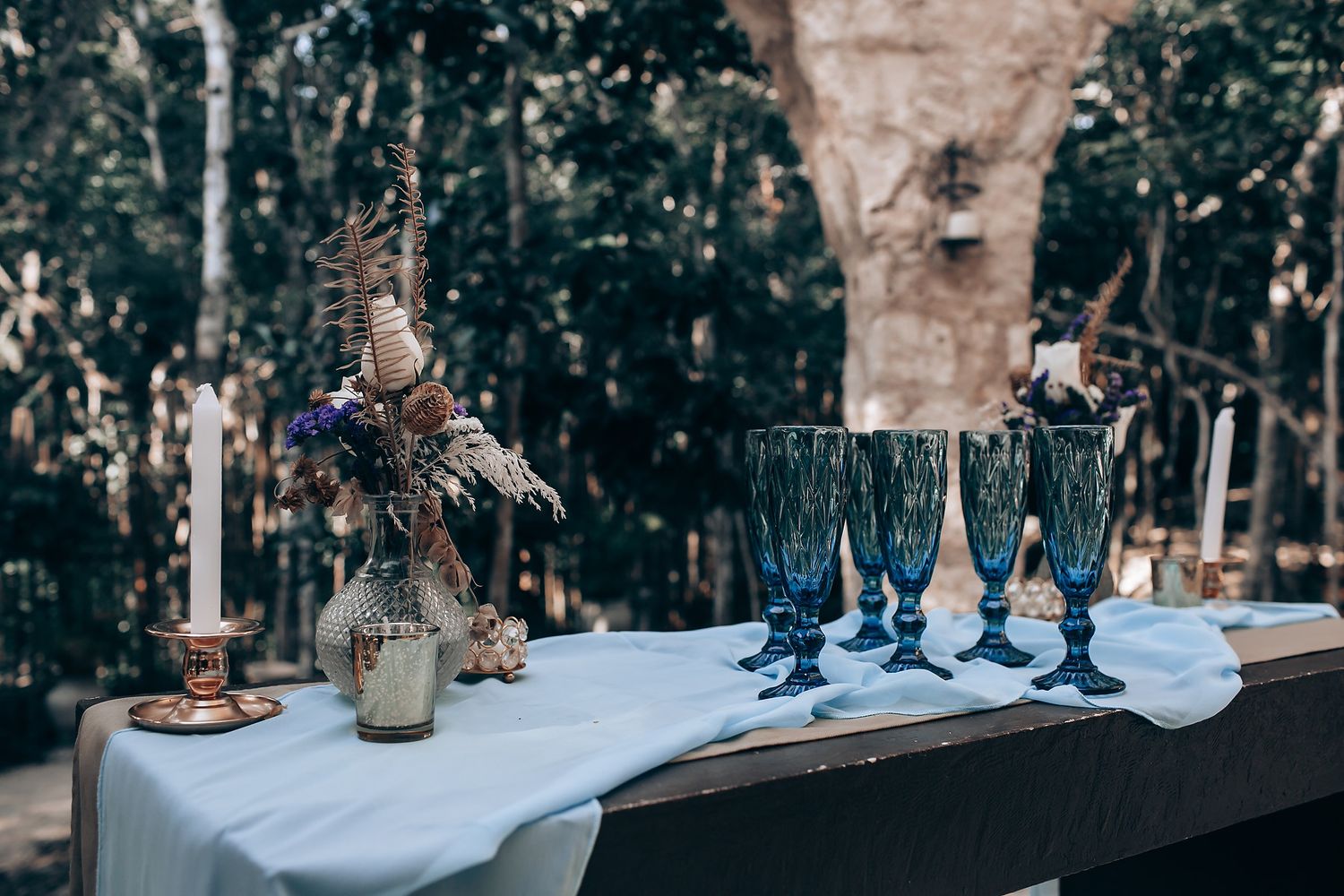 A rustic outdoor wedding reception table setting with vintage glassware and natural decor in a wooded area.