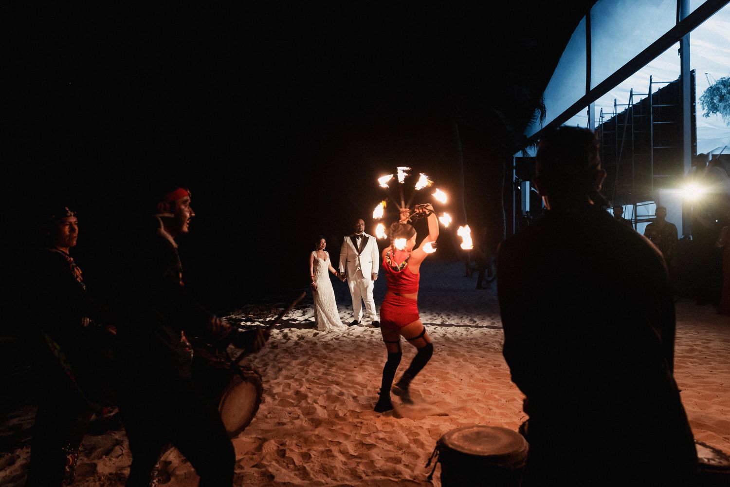 Fire dancer in red performs on sandy beach at night during wedding celebration with couple watching.