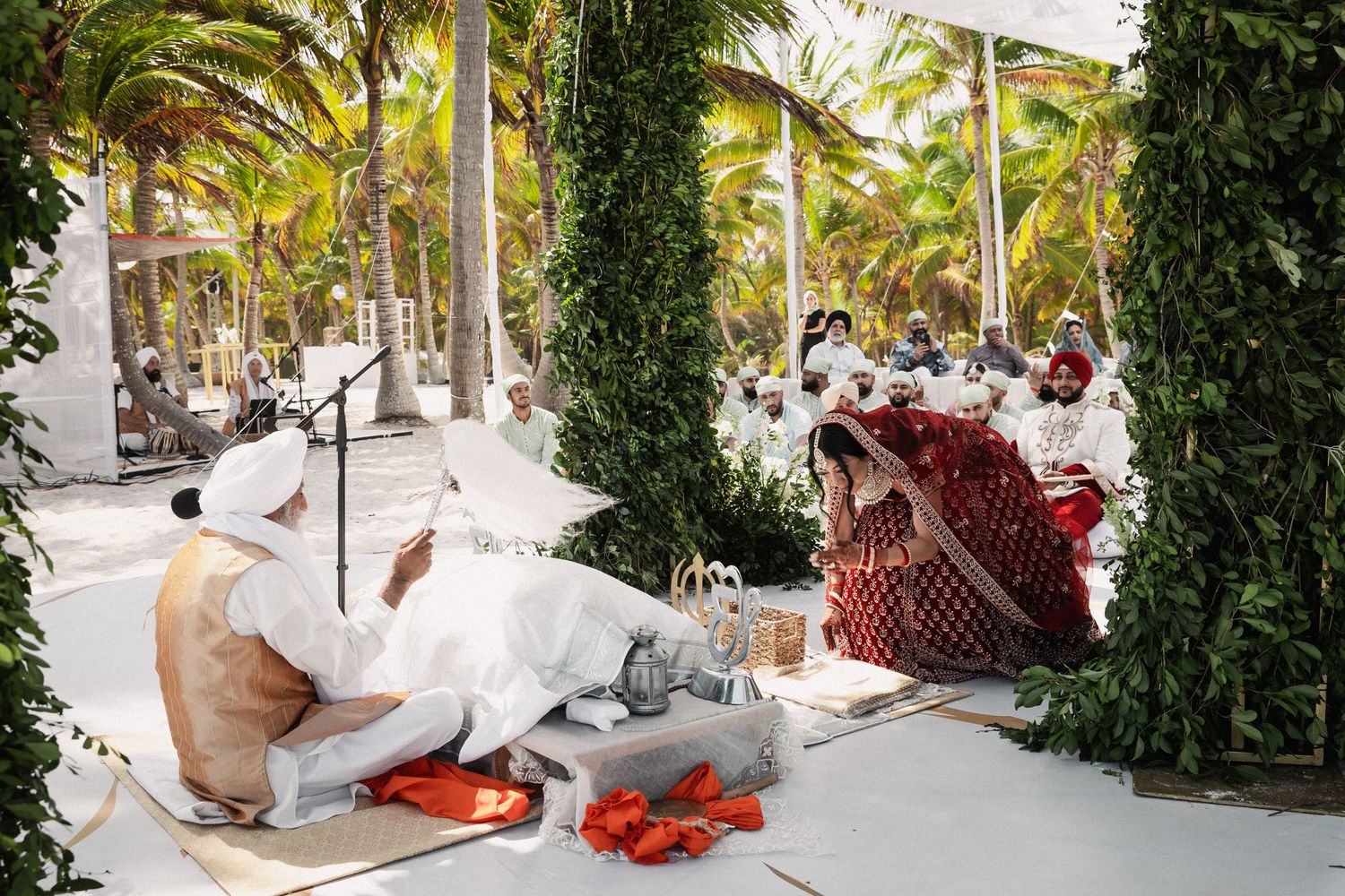 Sikh wedding ceremony outdoors with priest reading scripture beneath a lush green arch adorned with red rose petals.