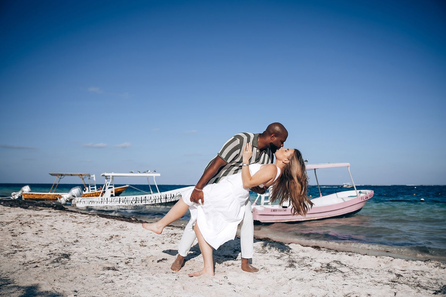 Interracial couple sharing a romantic dip kiss on a sandy beach with colorful boats behind them.