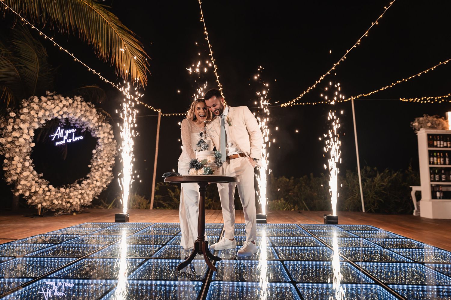 Bride and groom cut wedding cake on illuminated dance floor surrounded by sparkling fireworks at night.