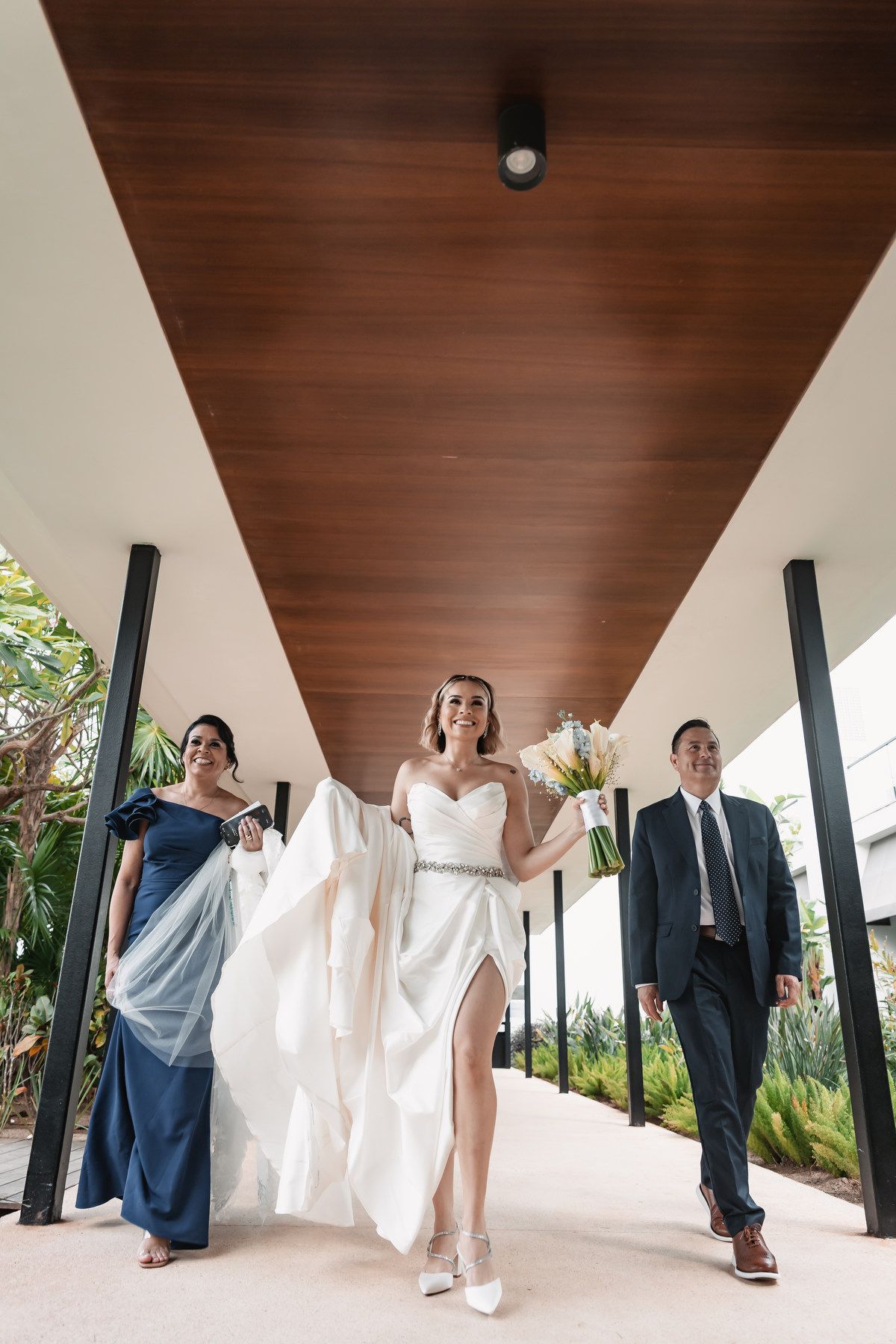 Bride in white gown walks with parents down modern outdoor corridor holding white bouquet.