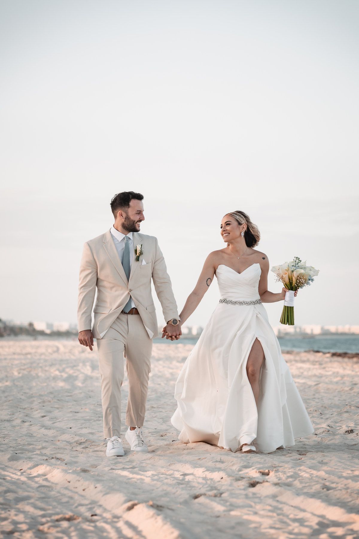 Newlyweds walking hand in hand on beach at sunset.