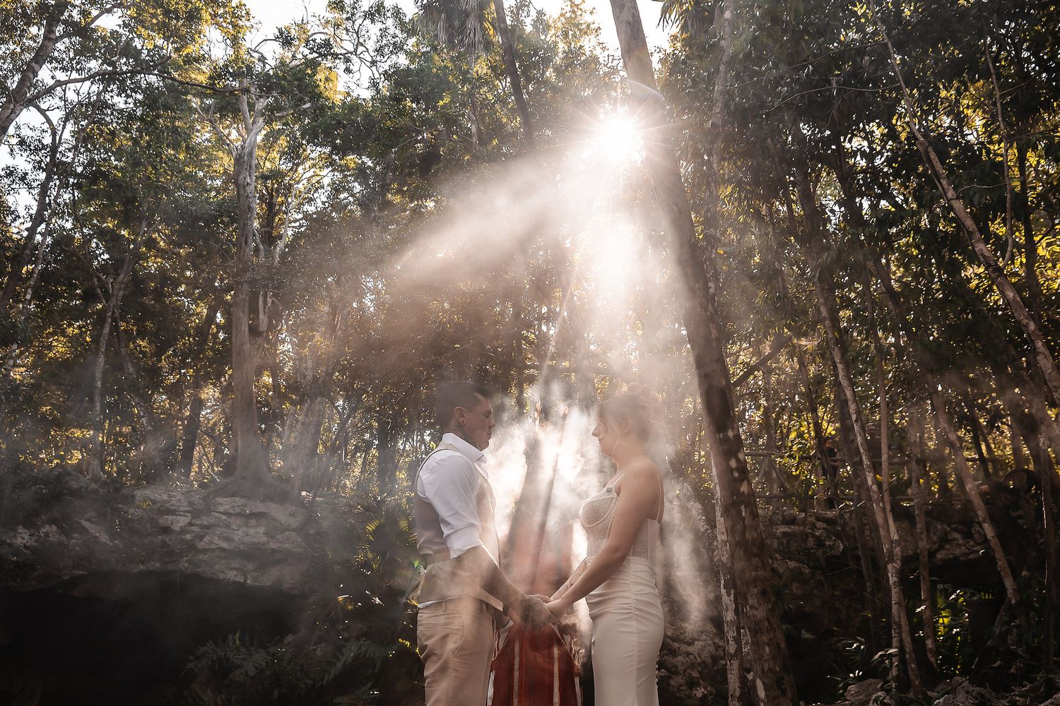 Couple embracing in a sunlit forest with dramatic light rays filtering through trees.