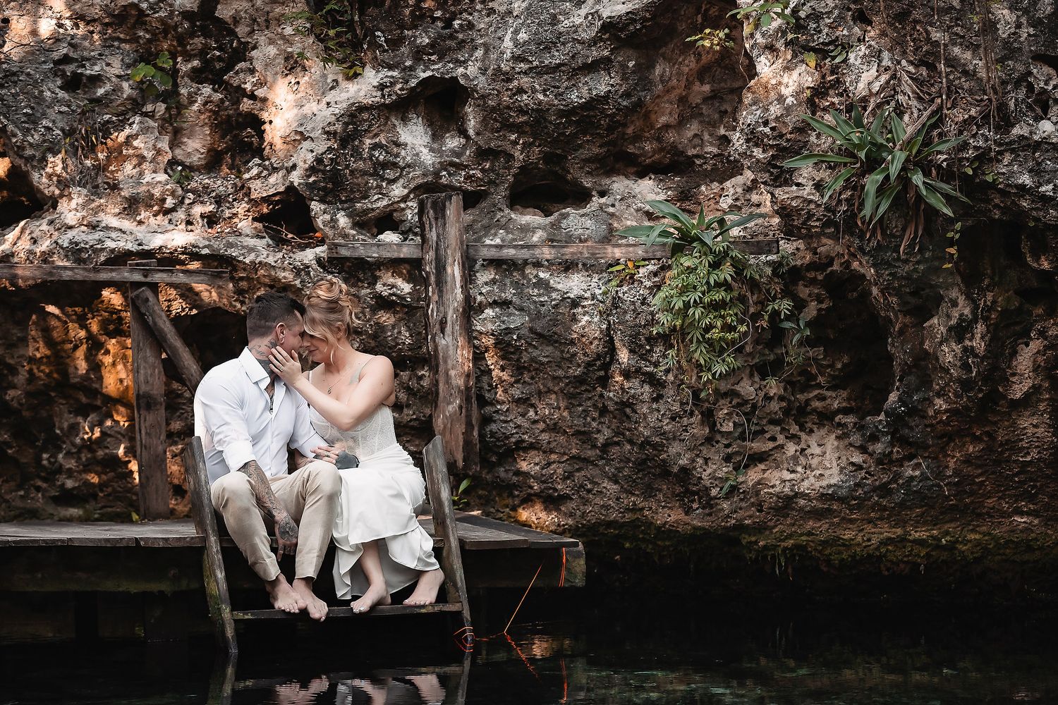Bride and groom share a romantic kiss on a wooden dock beside a serene cenote surrounded by ancient rock walls.