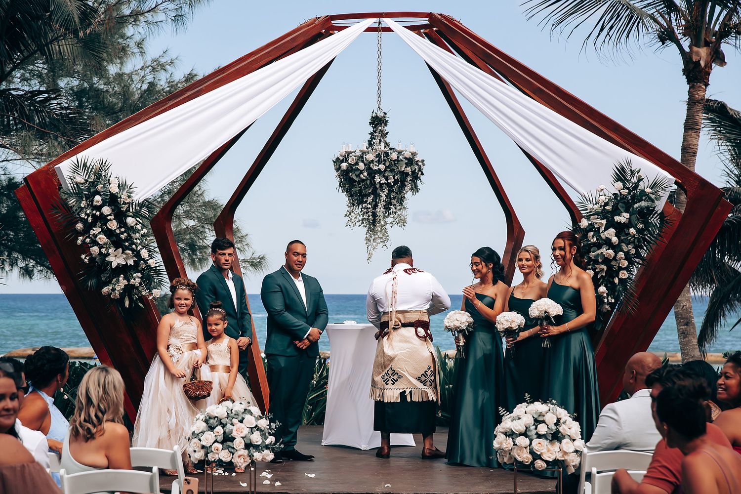 Outdoor wedding ceremony under a red and white draped arch with floral arrangements and ocean backdrop.