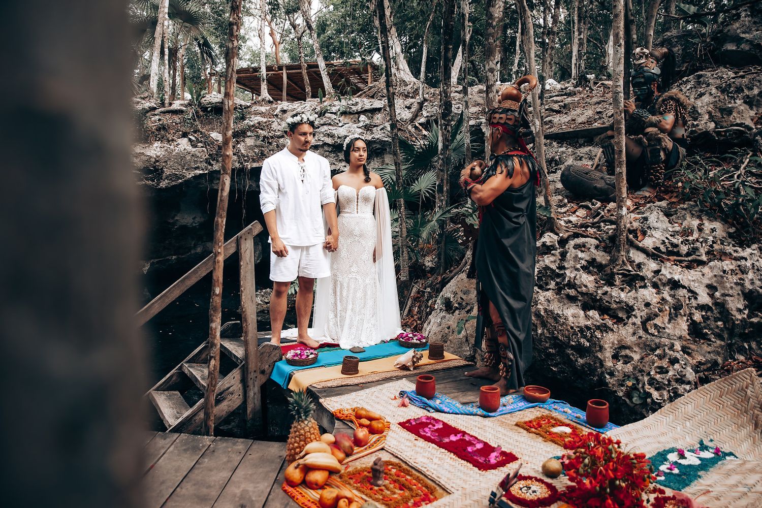 Tropical beachside picnic setup with white blankets, pillows and fresh fruits arranged on wooden platforms.