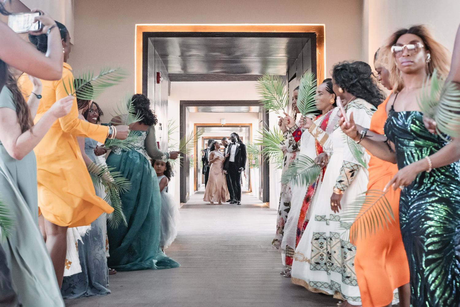 Wedding guests in vibrant green and orange dresses lining a hallway for a celebratory processional.