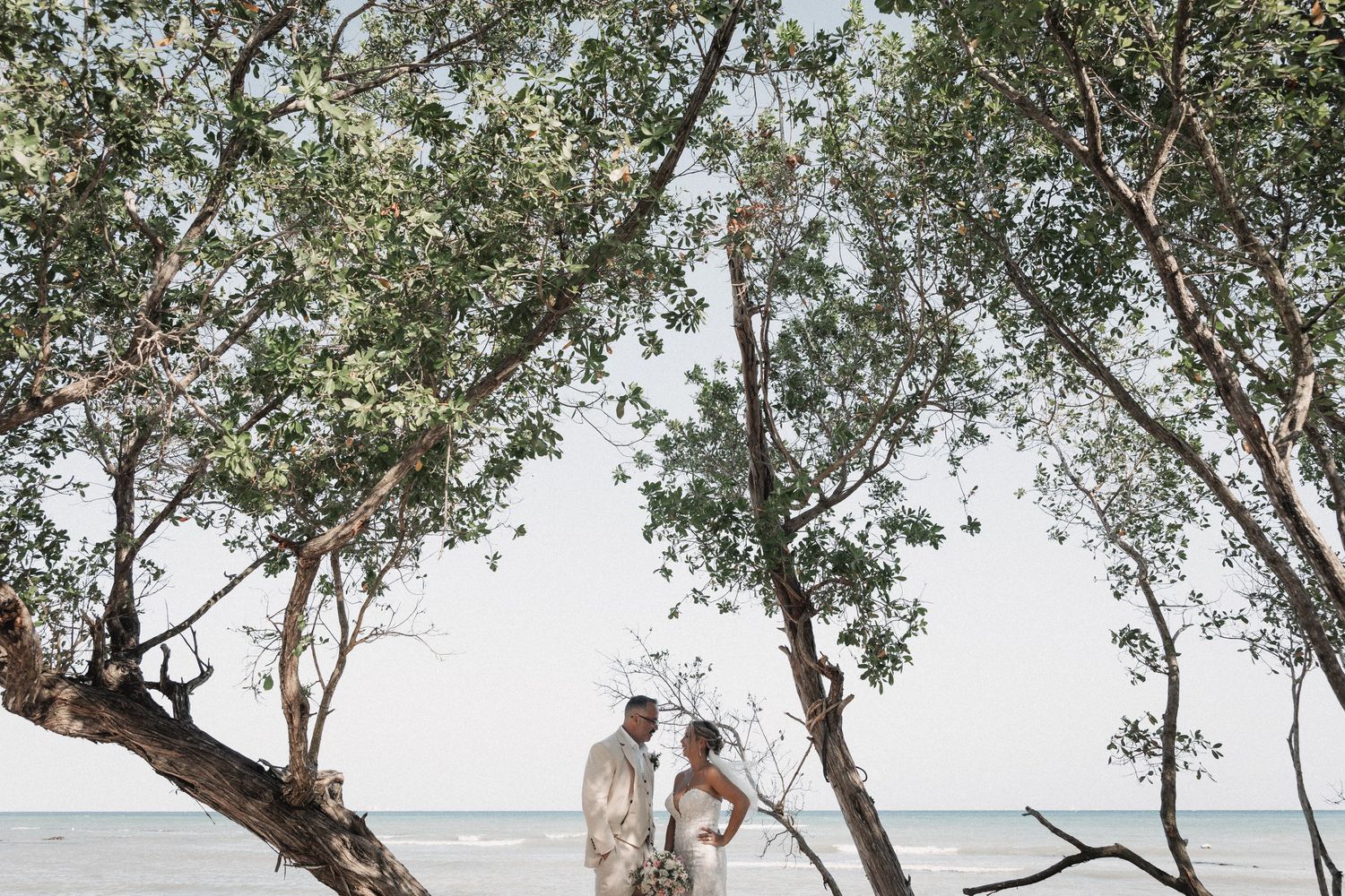 Bride and groom stand romantically between tropical trees with ocean view on their wedding day.