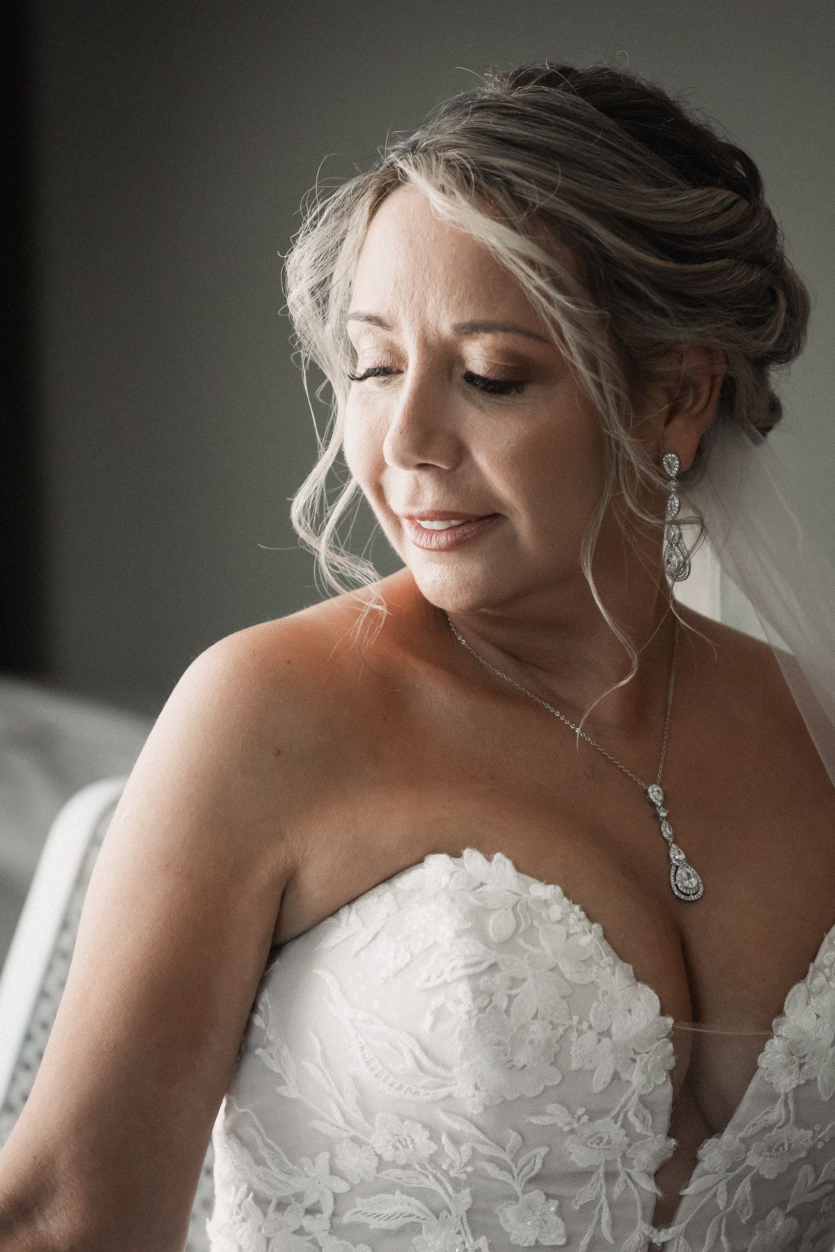 Bride in strapless lace wedding gown with crystal drop earrings and necklace, eyes closed, smiling softly.