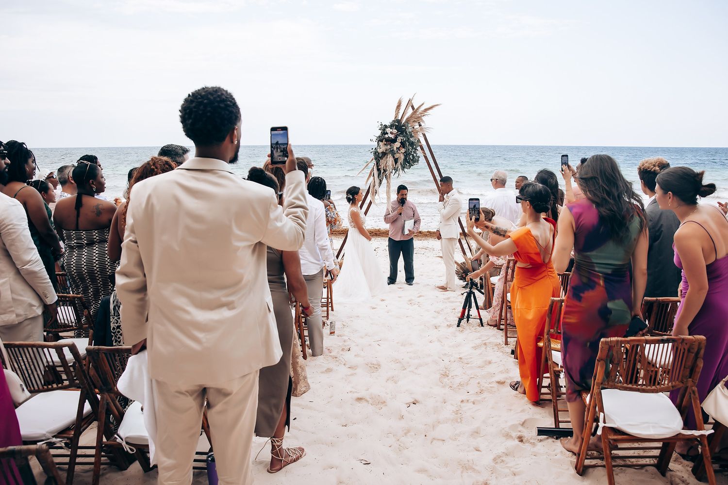 Beach wedding celebration with guests gathered on a sunny tropical shoreline.