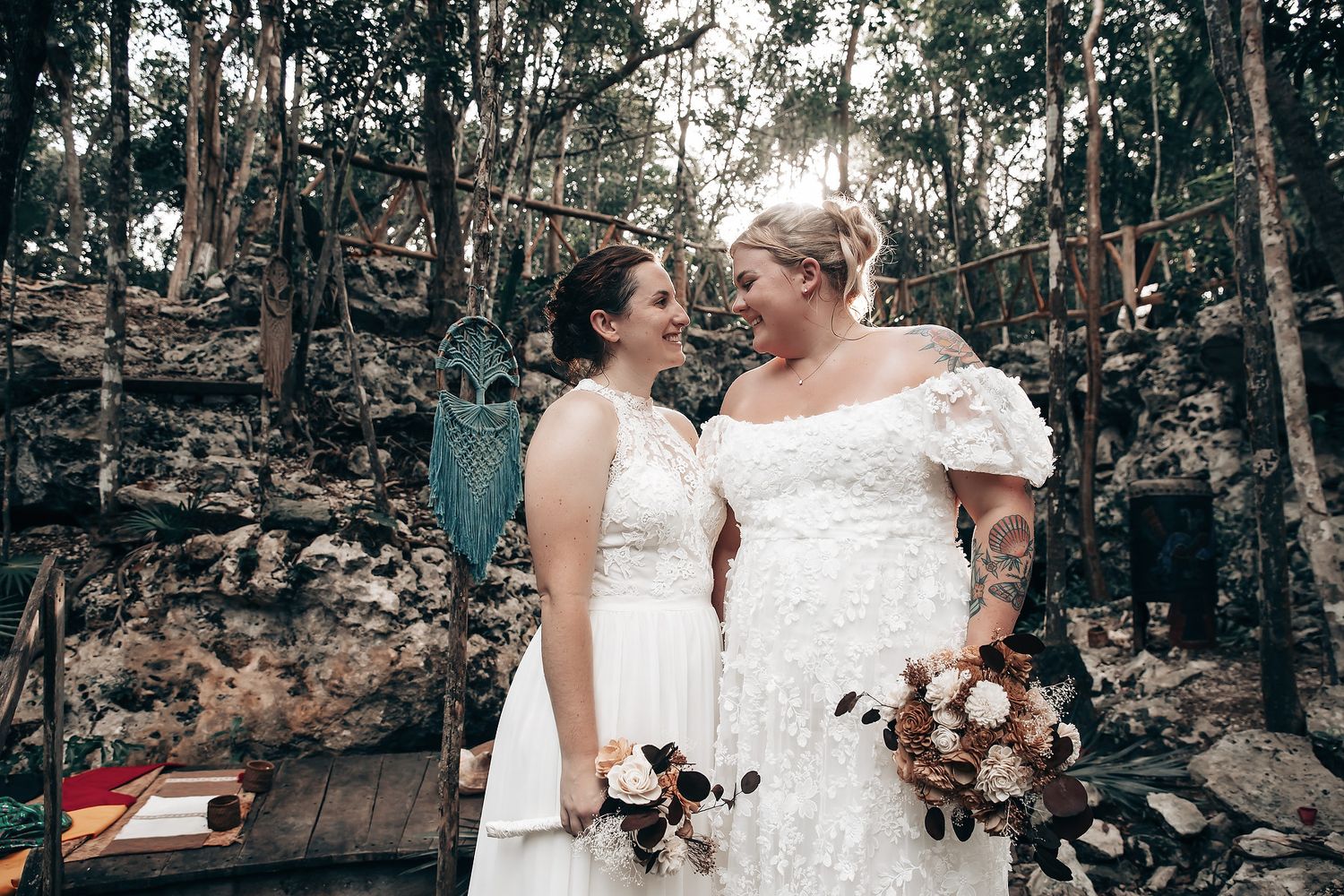 Two brides in white wedding dresses embrace in a lush, forested environment.