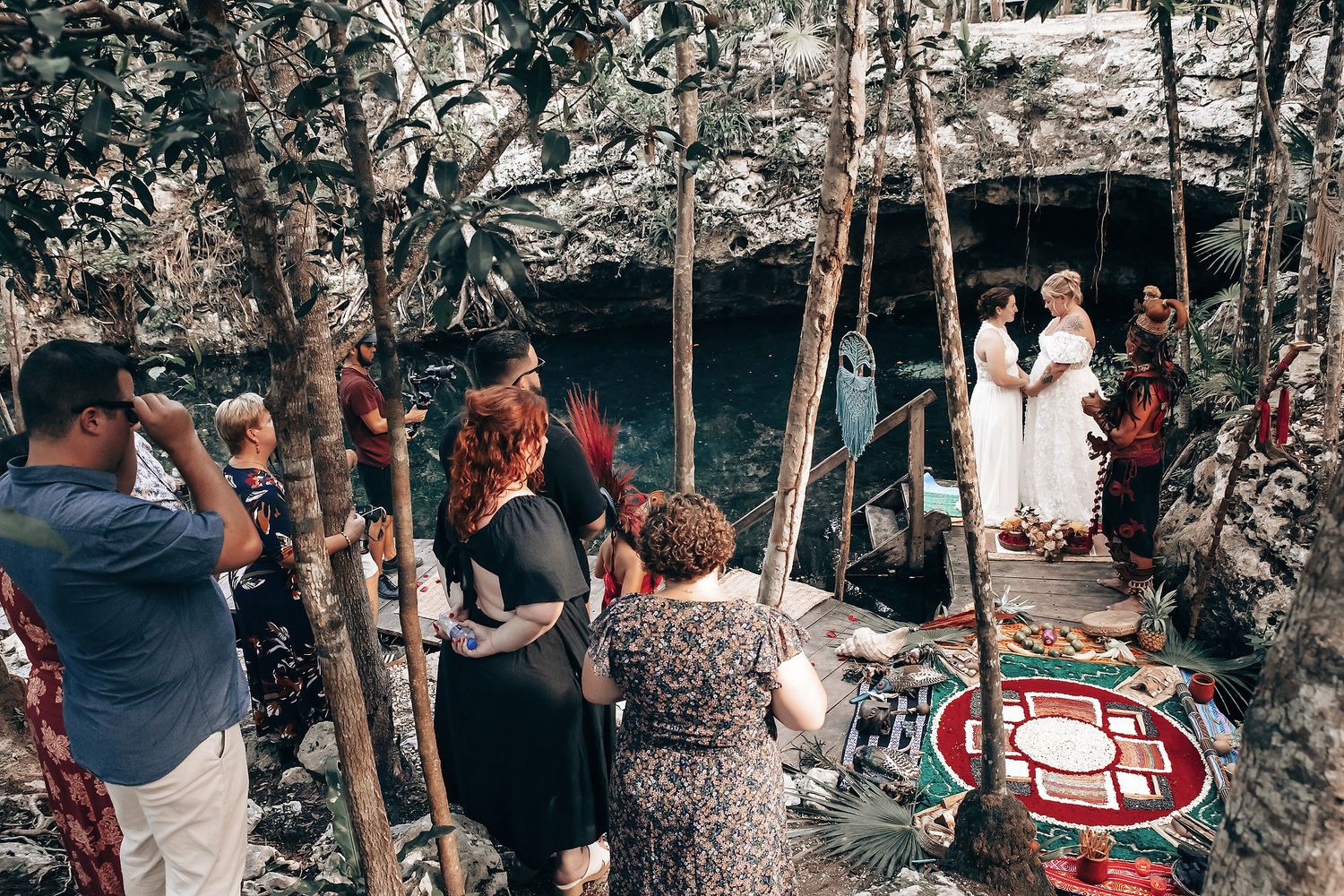A group of people in traditional cultural attire gather around a couple in a wedding ceremony in a wooded area.