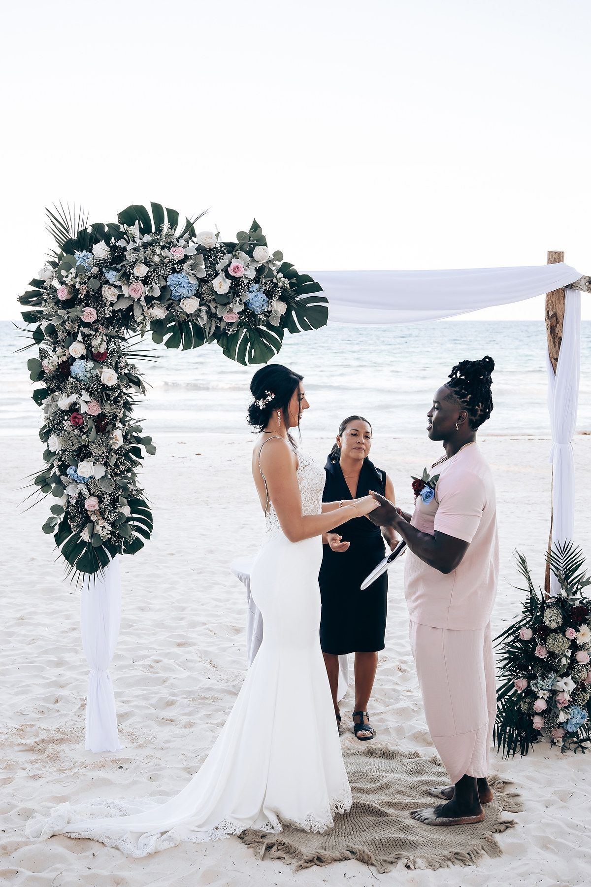 A wedding ceremony takes place on a beach, with a floral arch and two people standing at the altar.