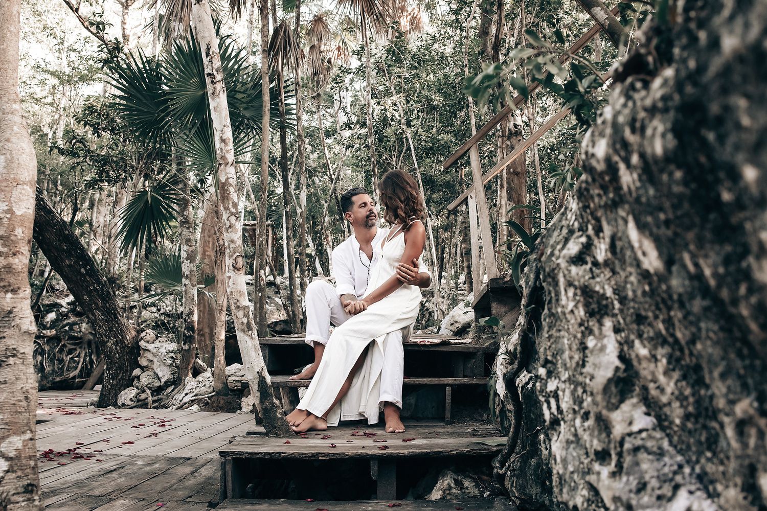 A couple stands together in a tropical forest setting, surrounded by lush vegetation and decorations.