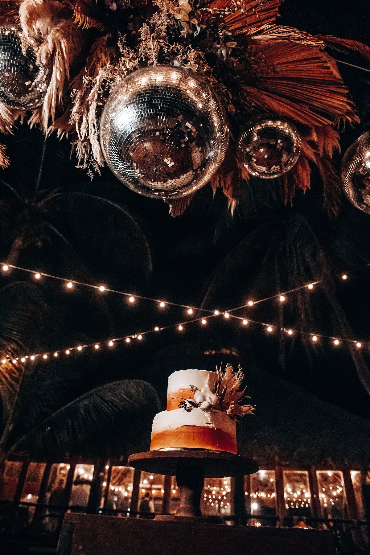 Wedding cake displayed under disco balls and string lights with feather decorations.