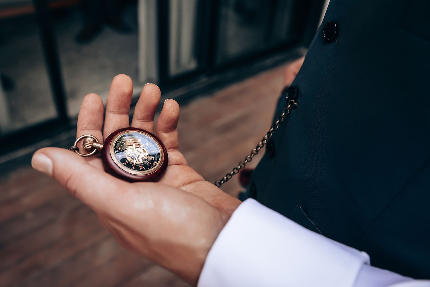 A vintage pocket watch with a chain is held up against a blurred background, showing its antique design and metallic details.