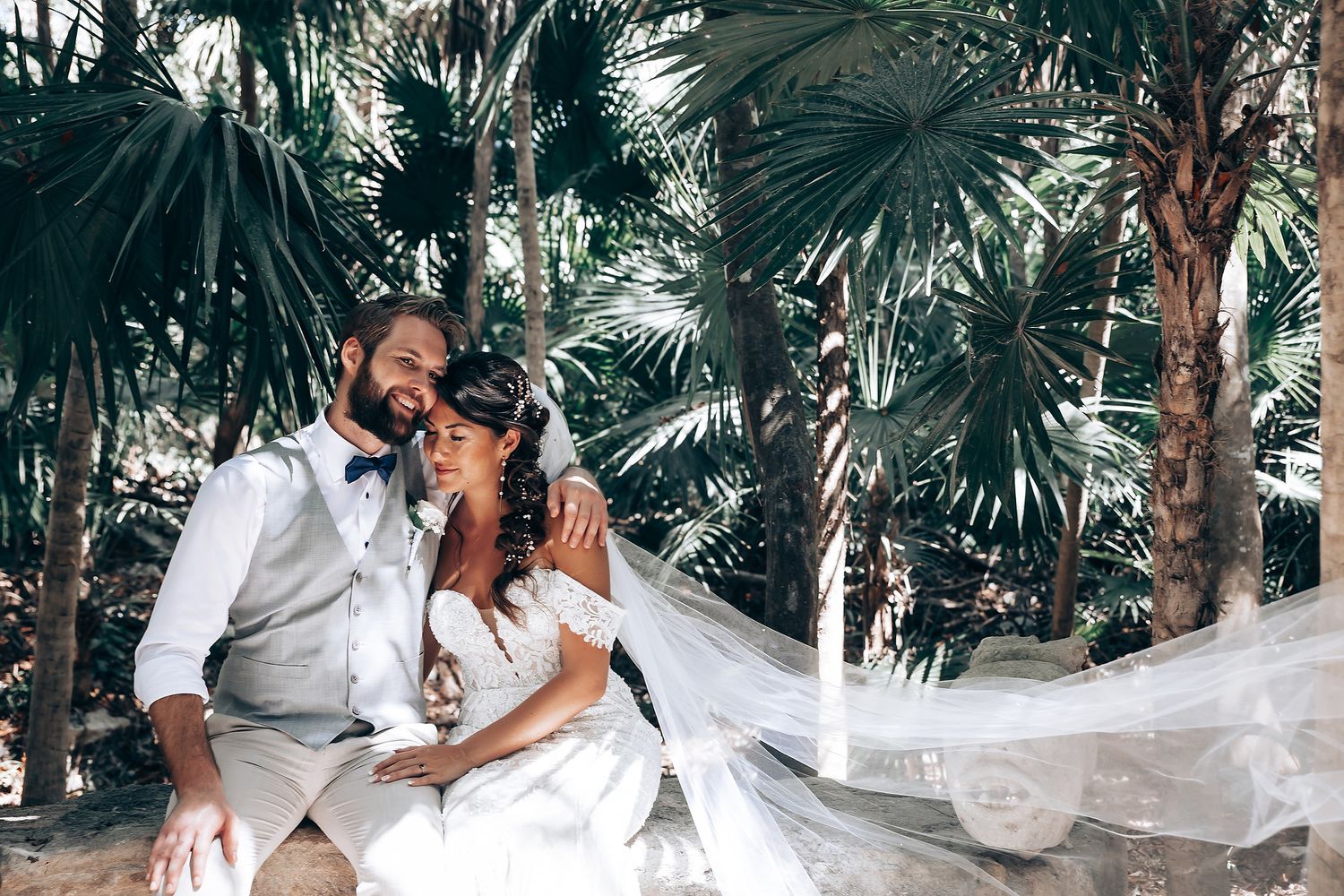 Newlyweds share an intimate moment on a palm-lined beach path as bride's veil flows in the breeze.