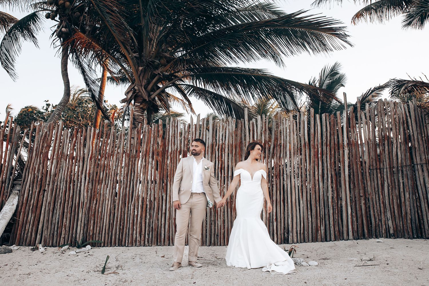 Wedding couple poses on sandy beach with palm trees and bamboo fence backdrop in natural lighting.