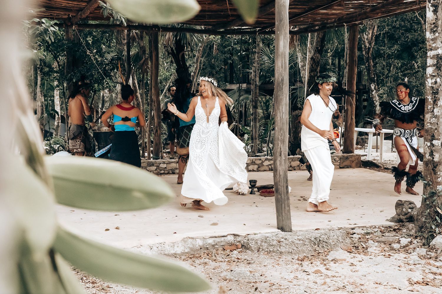 People in white clothing walk along a tropical path with lush greenery and trees creating a natural canopy overhead.