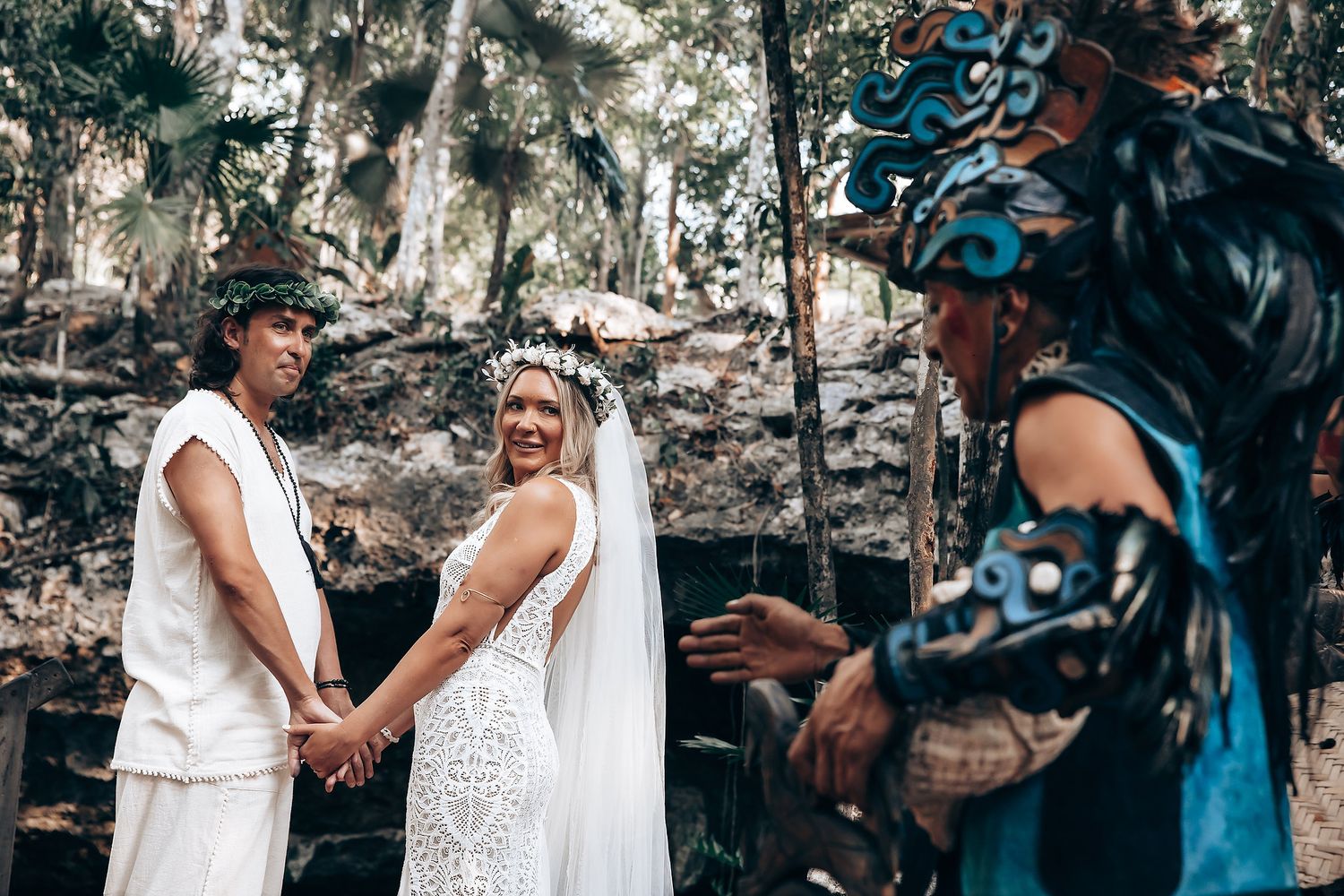 A couple in white outfits laugh and hold hands while seated against a stone wall backdrop.