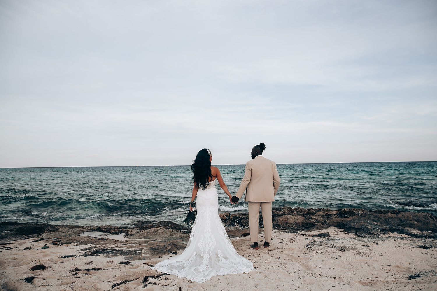 Couple stands at ocean's edge in wedding attire looking out at horizon.