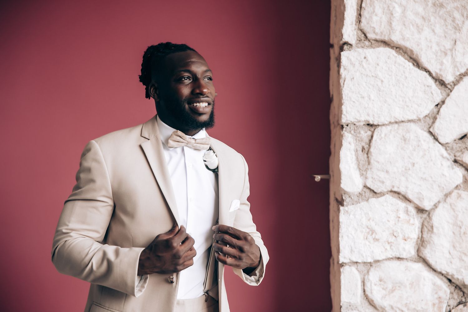 Groom in a tan suit poses against a burgundy wall with white stone accents.