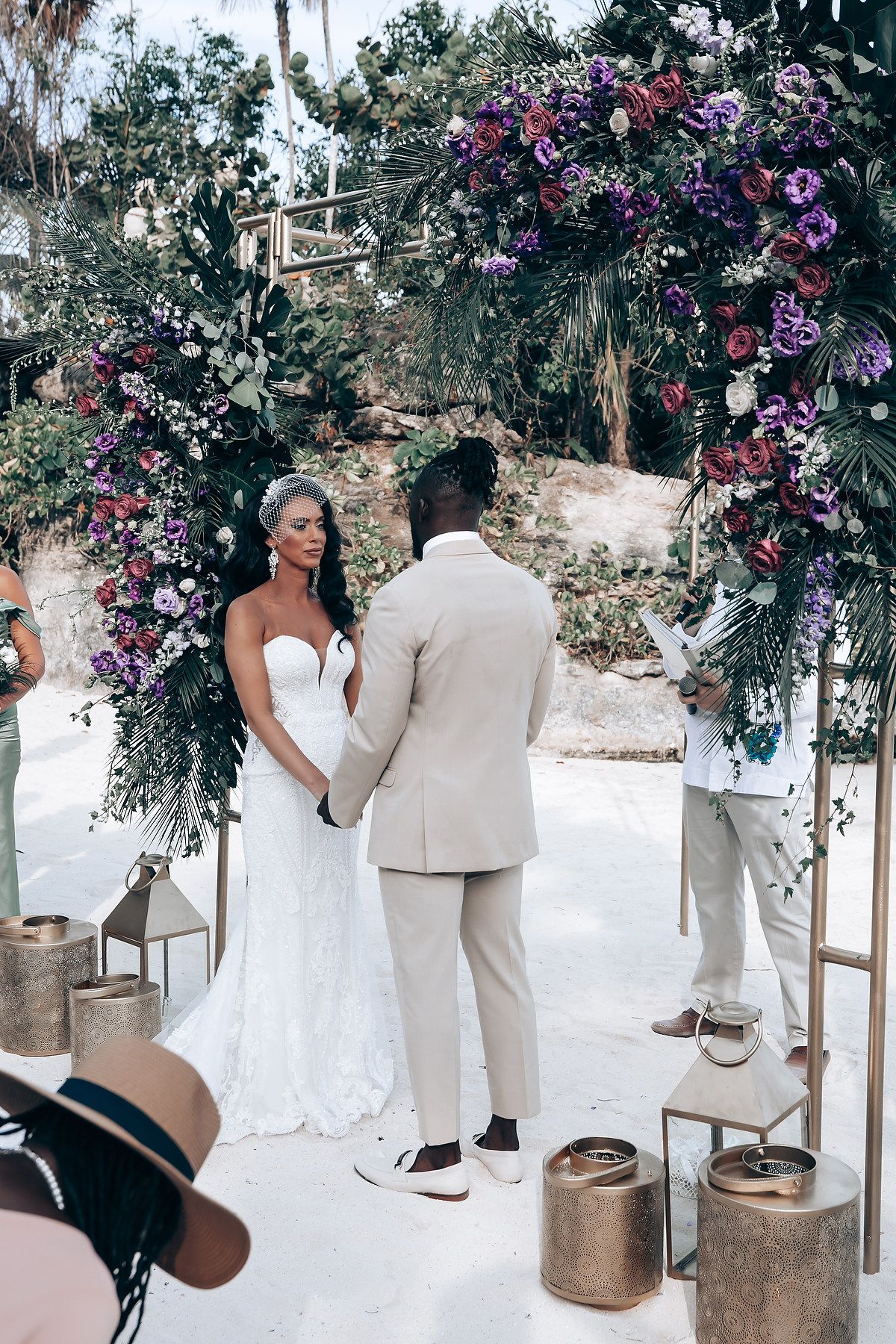 Wedding ceremony setup with floral arch and white chairs decorated with purple flowers in a tropical setting.