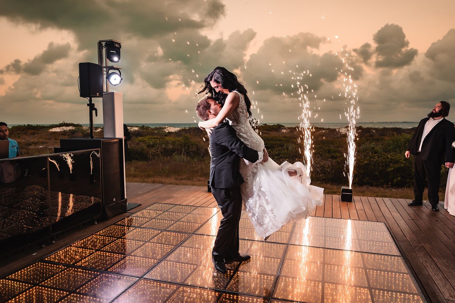 Groom lifts bride on outdoor dance floor with sparkling fireworks at a romantic beach wedding celebration at dusk.