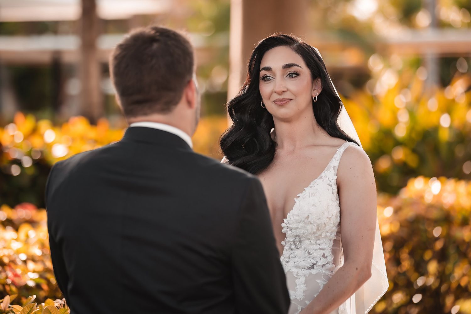 Bride in lace gown gazes at groom during outdoor wedding ceremony with colorful floral backdrop.