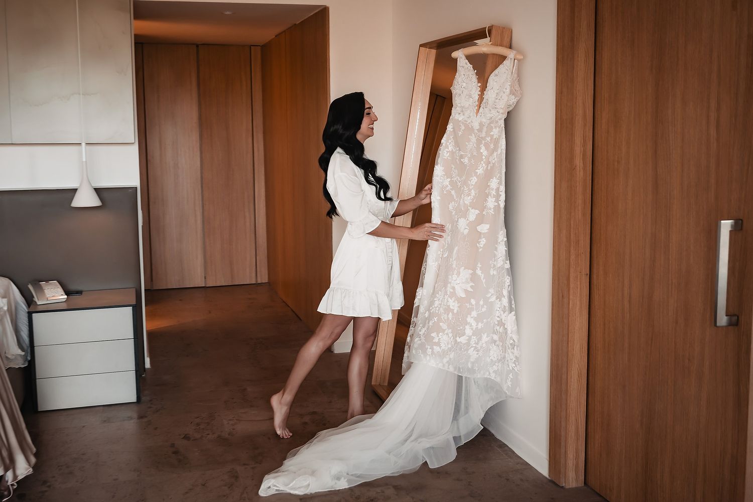 Bride in white robe admiring her floral lace wedding dress hanging in a hotel room.