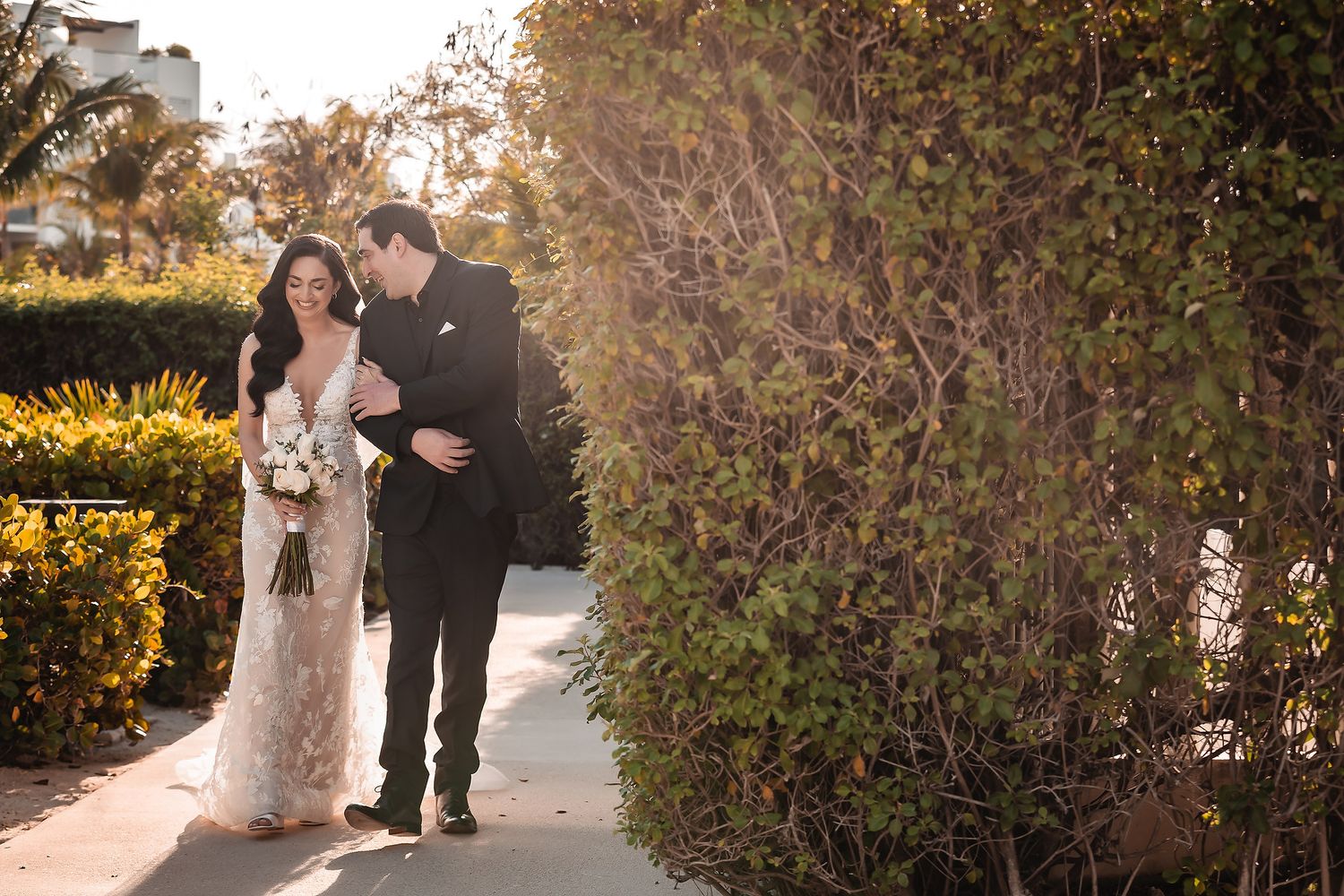 Bride and groom walking together on a sunny outdoor path, surrounded by lush green hedges on their wedding day.
