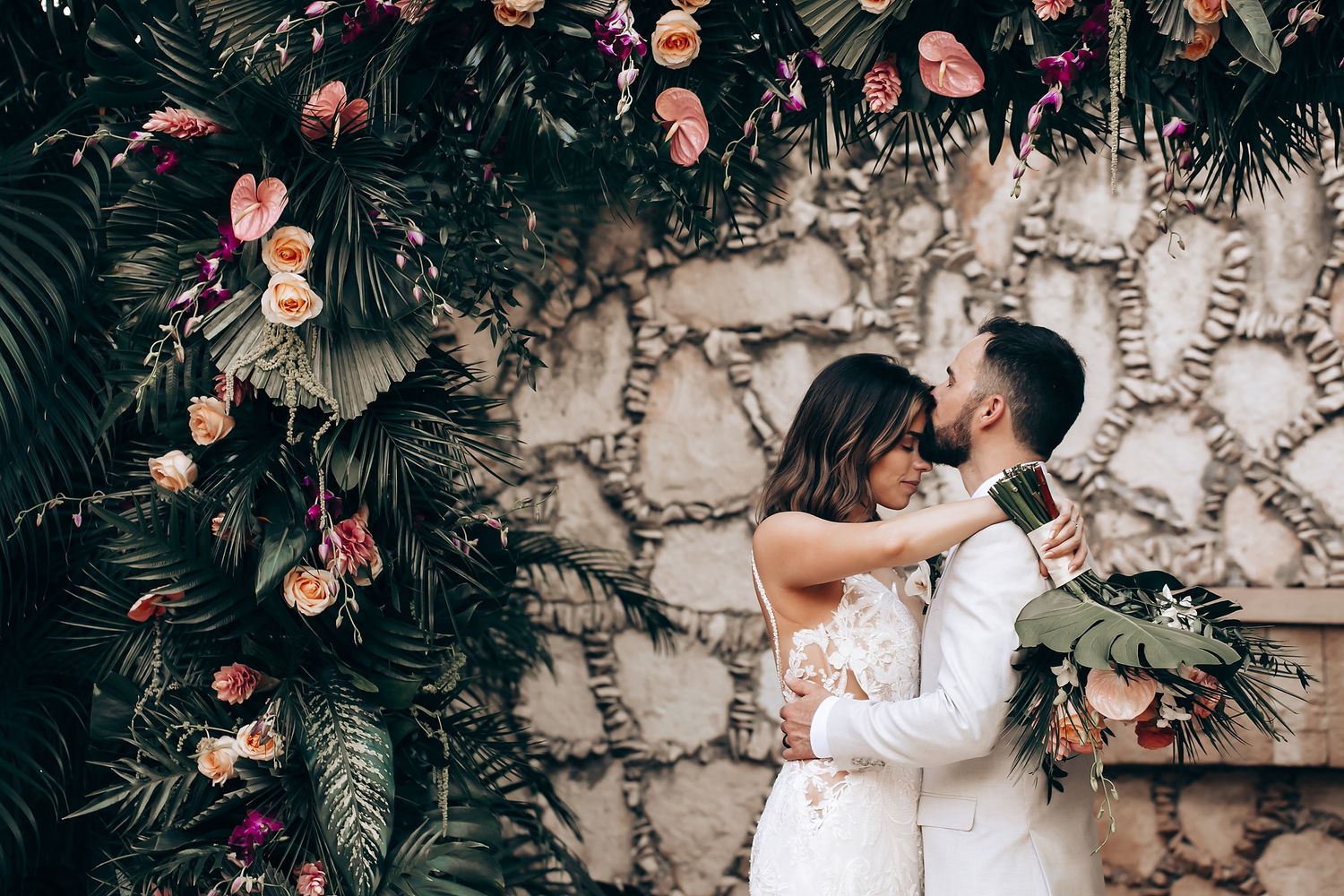 A couple embraces against a stone wall adorned with pink flowers and lush greenery.