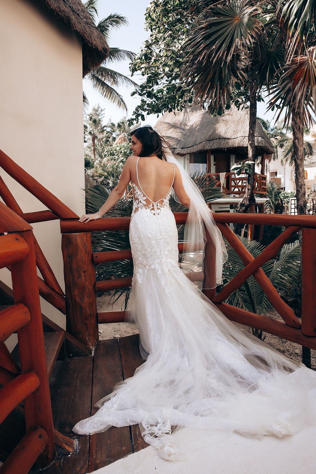 A flowing white lace wedding dress with a long train displayed on a wooden balcony with tropical palm trees in background.