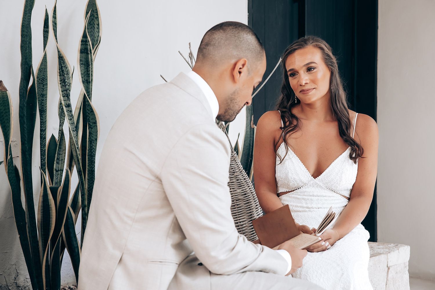 A romantic couple in white outfits embraces against a modern interior wall with vertical plants.