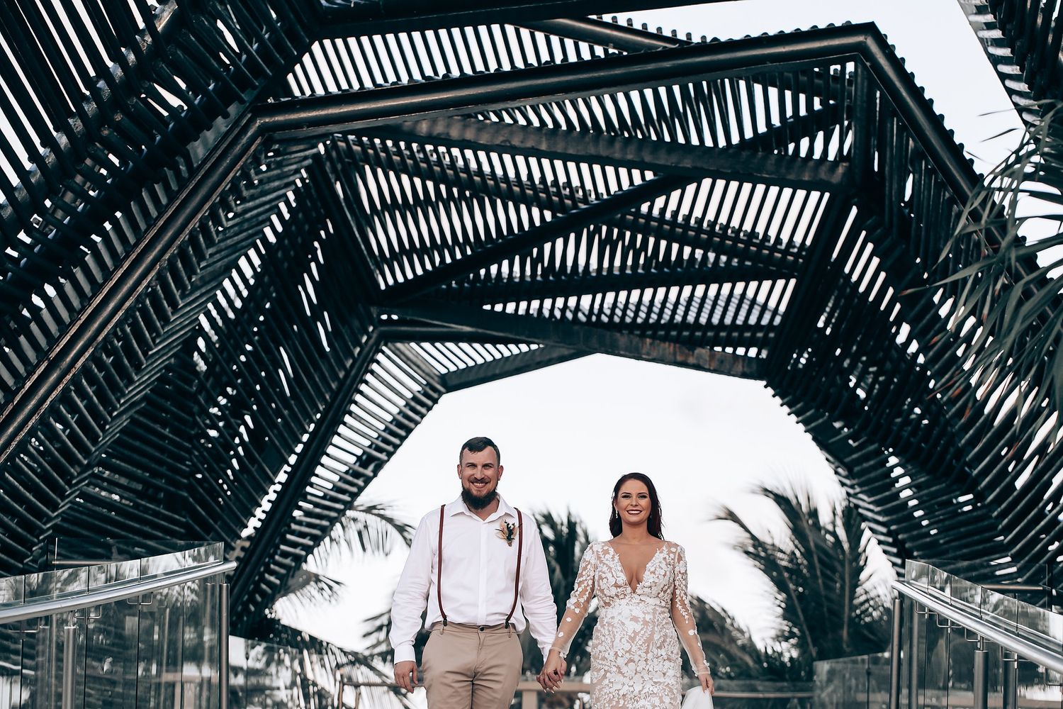 Newlyweds walking together under modern architectural structure with palm trees visible in background.