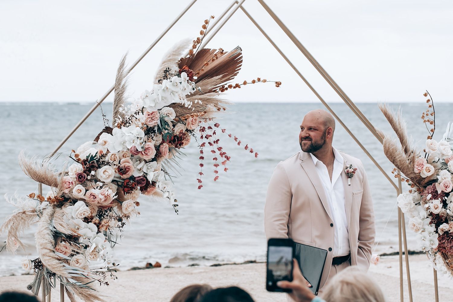 Beach wedding ceremony setup with geometric arch decorated in dried flowers and neutral tones.