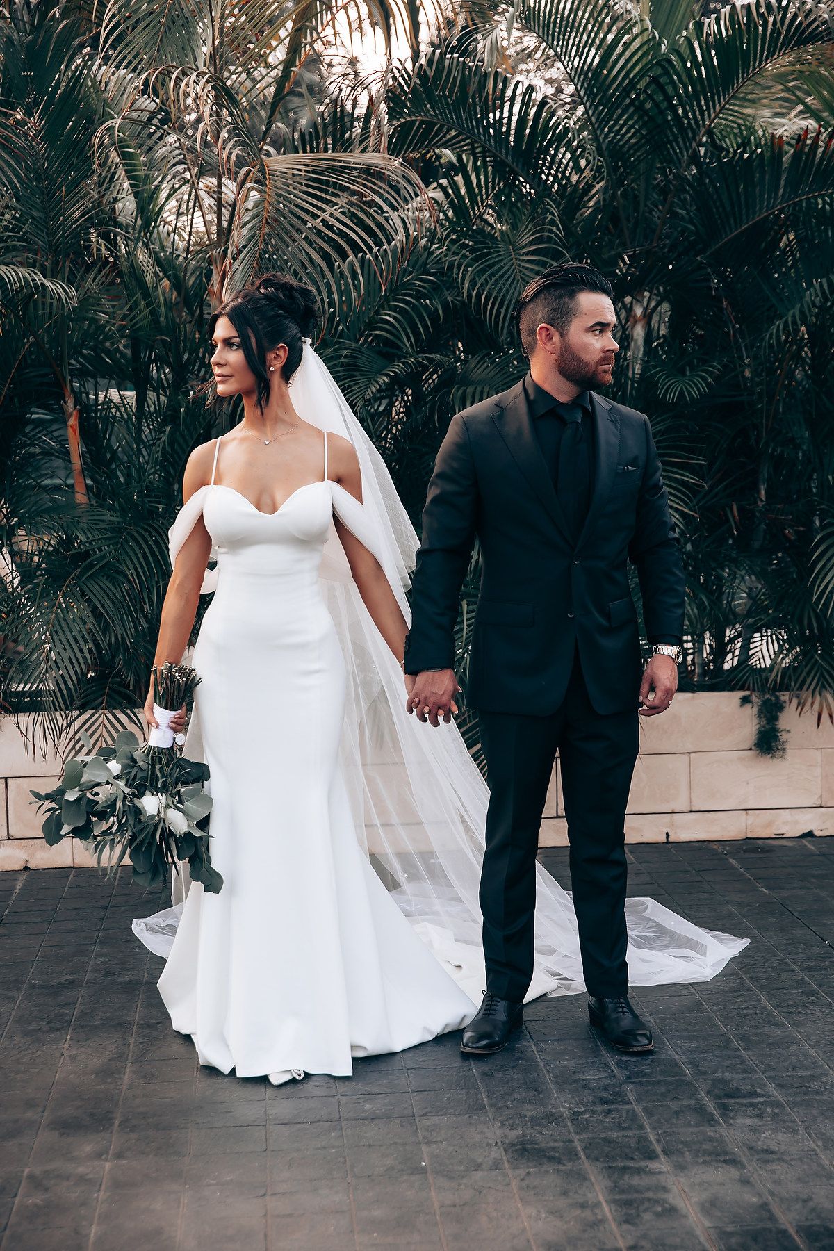 Bride in white gown and groom in black suit walking through tropical palm garden setting.