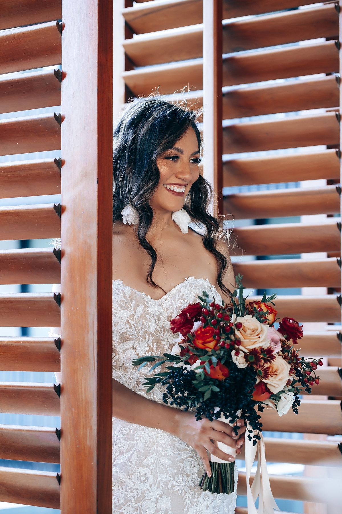 Elegant bride in lace dress holds vibrant red and blush floral bouquet against wooden shutters.